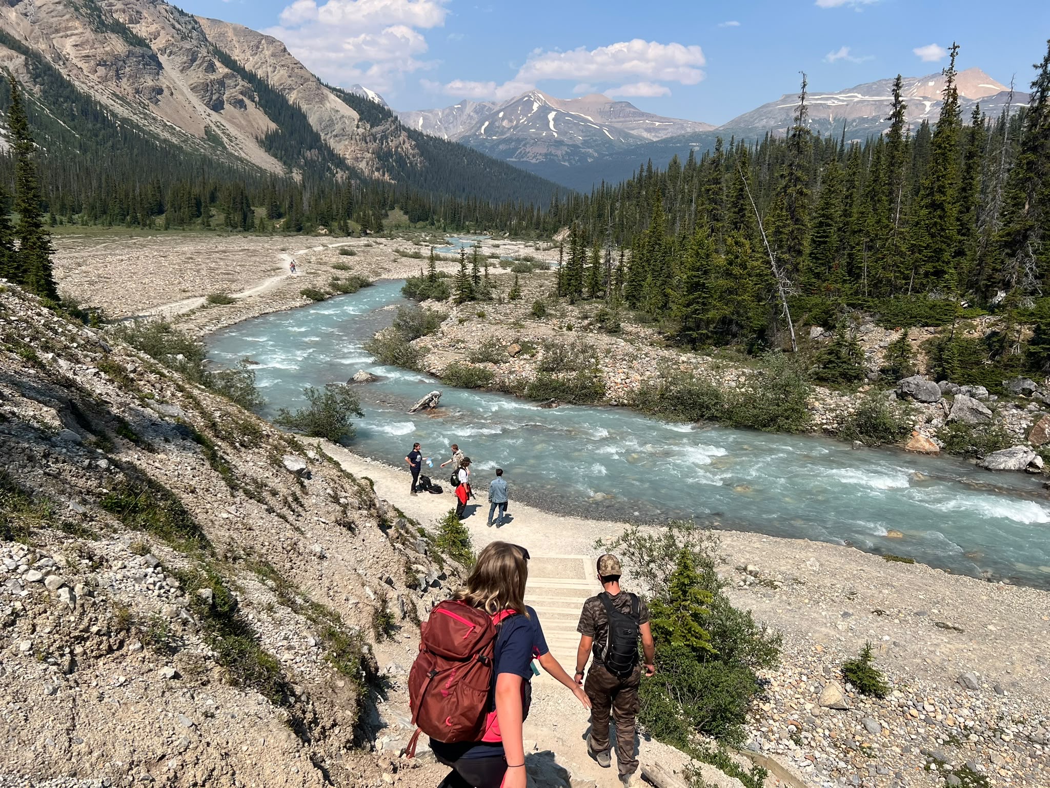 Mountain river / hiking Campers walking toward a turquoise mountain river surrounded by forest and Rocky Mountain peaks.